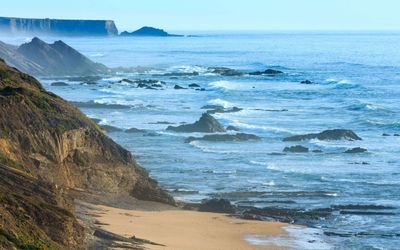 View along the wild Costa Vicentina coastline with waves breaking over scattered rocks and layered cliffs fading into the Atlantic haze