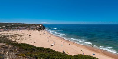 Elevated view of Praia de Monte Clérigo with beachgoers on golden sand, Atlantic waves, and the village on the hillside behind