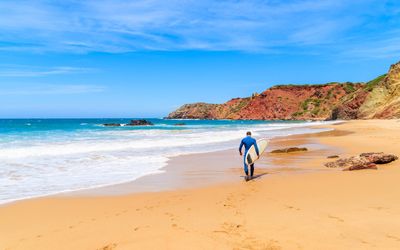 Surfer in a blue wetsuit carrying a surfboard walking towards the waves on golden sand with red cliffs in the background
