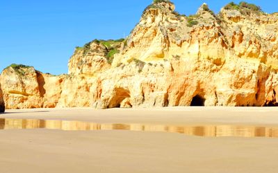 Dramatic golden-orange limestone cliffs with cave openings at the base, wet sand reflecting the rock formations