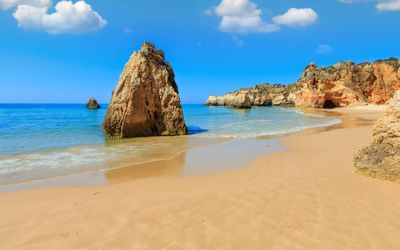 Golden sand beach with a tall sea stack in the shallows and orange-red cliffs with caves in the background