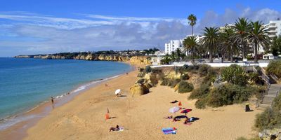 Wide golden beach with beachgoers under umbrellas, palm-lined promenade, and limestone cliffs in the distance