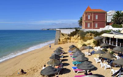 Beach section with straw parasols and sunbeds, distinctive red building on the clifftop, and beachside restaurant