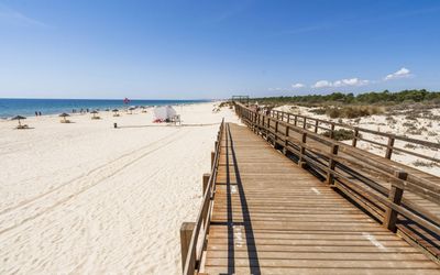 Wooden boardwalk running alongside the wide white sand beach with straw umbrellas and dunes on the right
