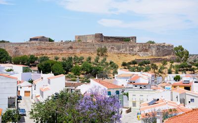 Castro Marim Castle on the hilltop seen from the town below with whitewashed houses, terracotta roofs, and a jacaranda tree in bloom