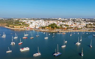 Aerial view of Alvor's harbour with dozens of sailboats and catamarans anchored in calm water and the village on the clifftop behind