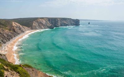 Arrifana beach's natural amphitheatre of towering cliffs with turquoise Atlantic waves and a distant sea stack