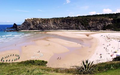 Praia de Odeceixe where the Seixe River curves around a sandbar to meet the Atlantic, with surf school and beachgoers on the golden sand